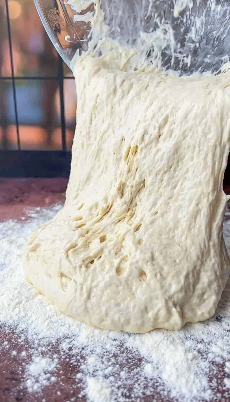 A close-up of sticky dough being poured from a bowl onto a floured surface. The dough has a light, airy texture, and the surface beneath is dusted with flour. A blurred background with hints of warm light is visible.