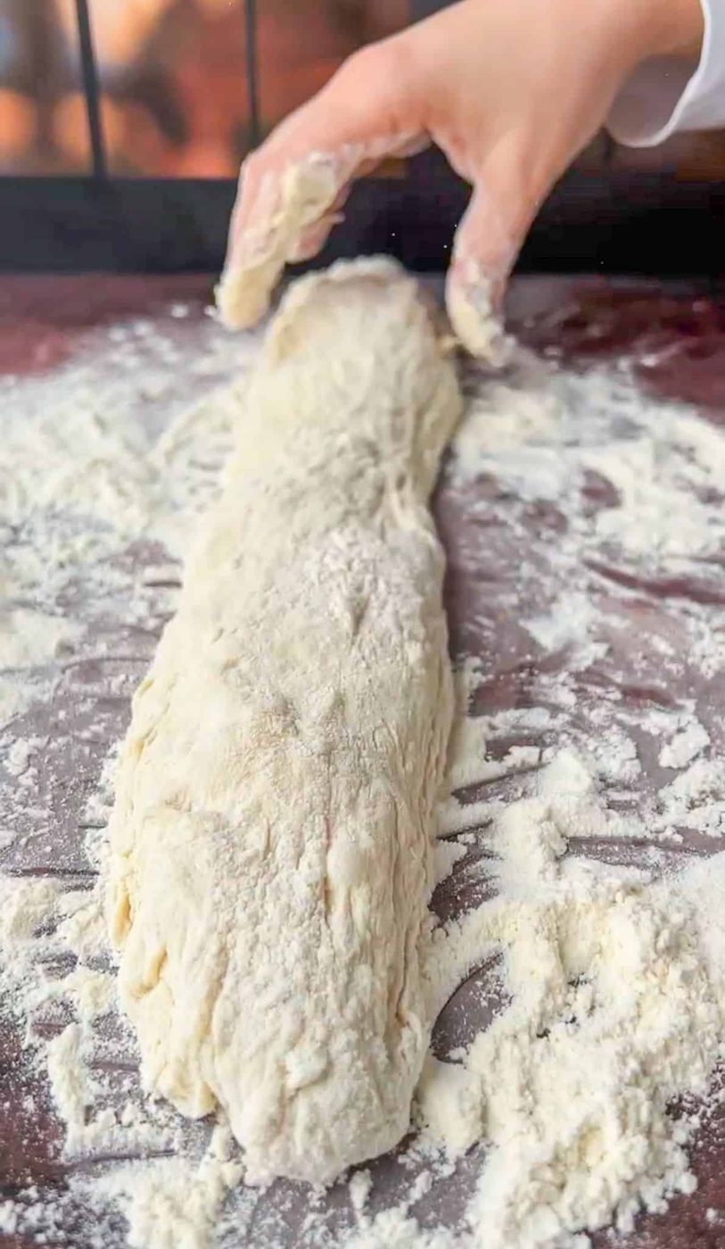 A person shaping a long piece of bread dough on a floured surface, with one hand gently folding the dough. The dough is lightly dusted with flour, and the surface is covered in flour as well.
