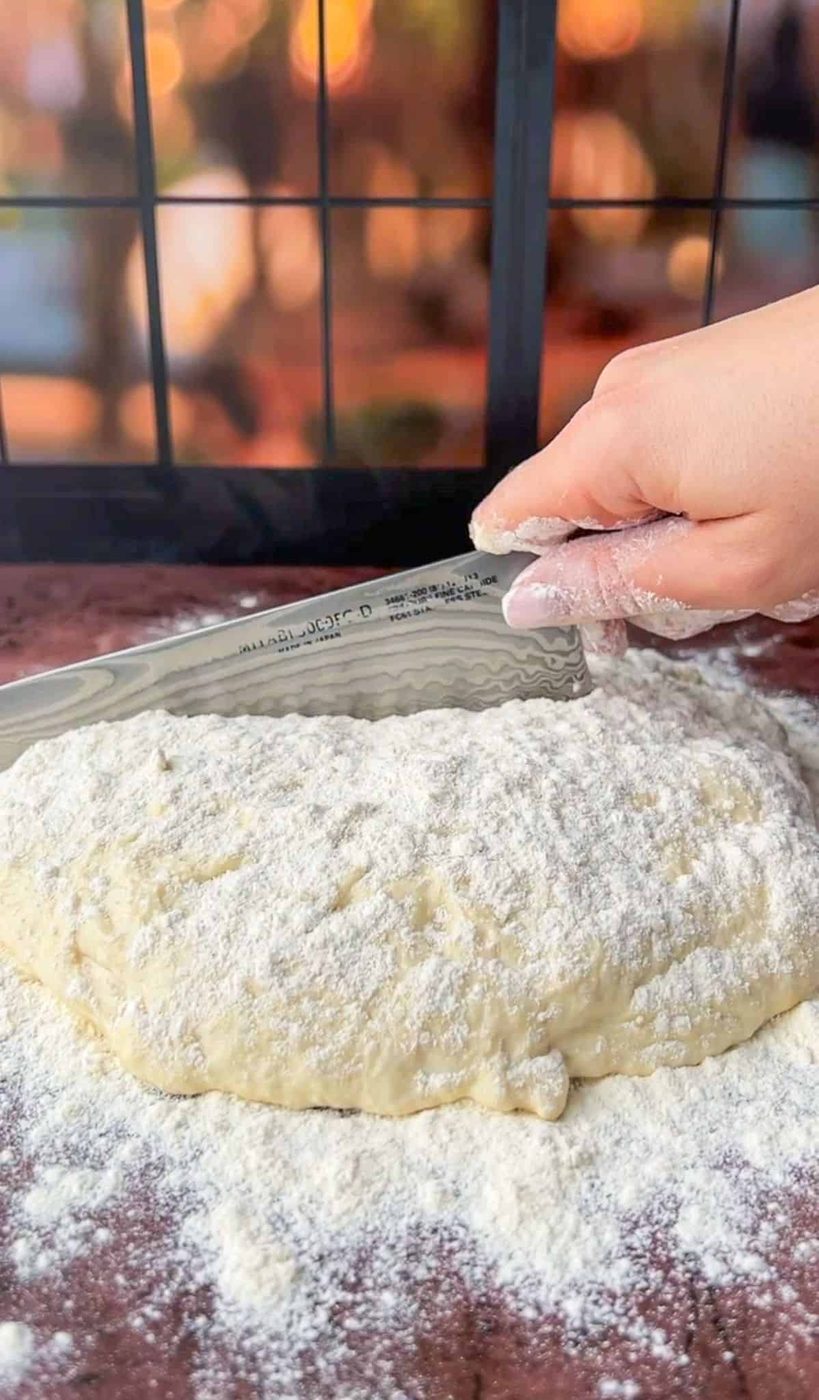 A close-up of a hand using a serrated knife to cut a floured dough on a brown surface. The dough is covered in a layer of flour. There is a blurred background with warm lighting.