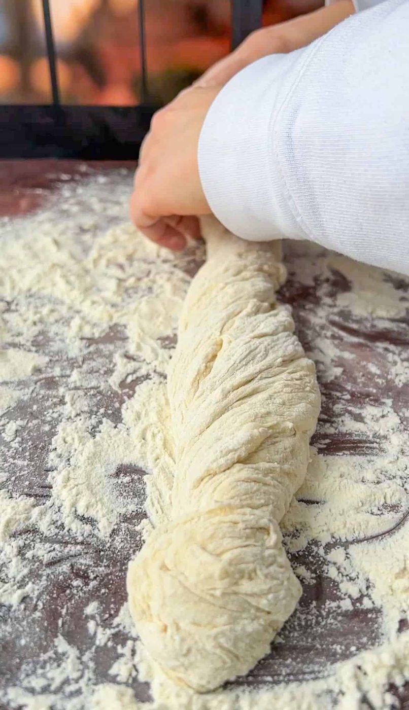 Person twisting a rope of dough on a floured surface, preparing bread or pastry. The dough is lightly dusted with flour, and the person's hand adjusts the twist.