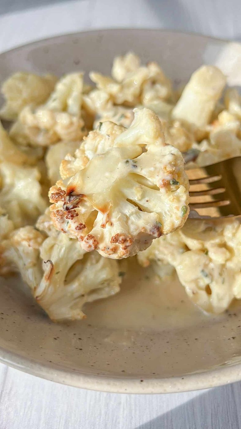 A fork is holding a piece of cauliflower, displaying a golden-brown edge, with sauce and herbs visible. The dish is set on a light-colored surface.