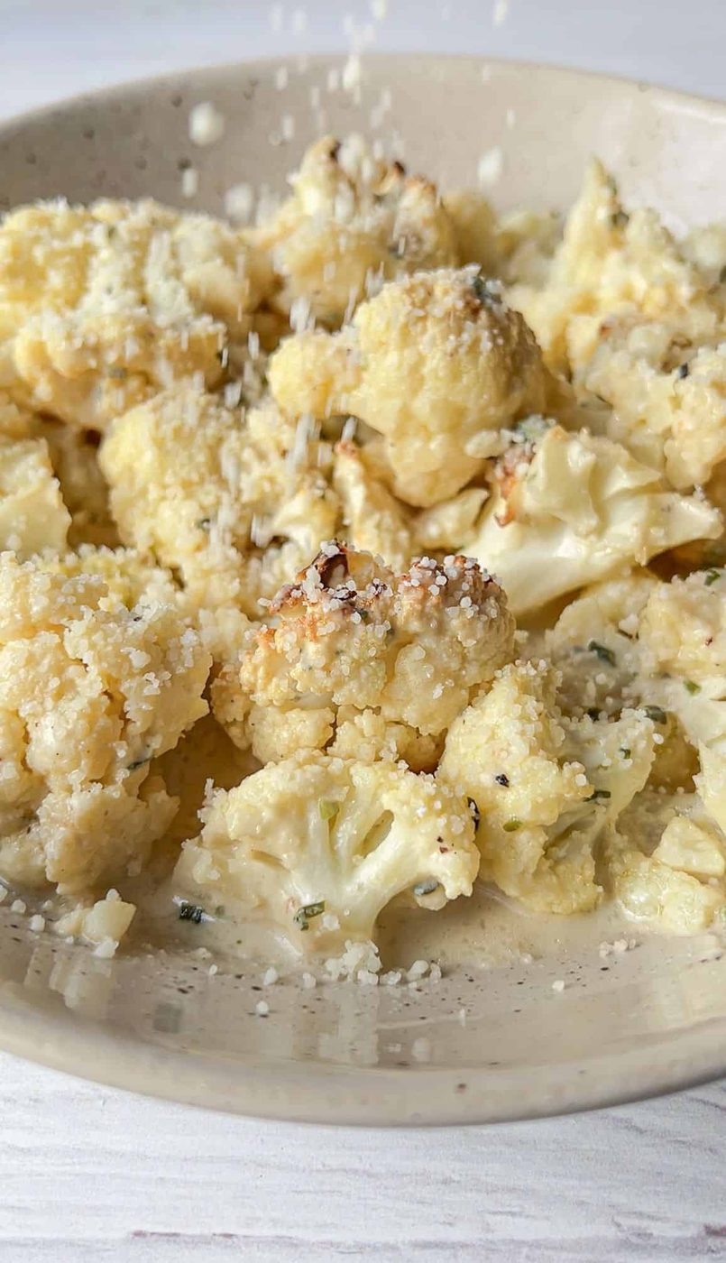 A close-up of the finished recipe sprinkled with grated cheese. The cauliflower is lightly browned and seasoned with herbs, visible in the creamy coating. The cheese is falling onto the vegetables, adding texture and flavor.