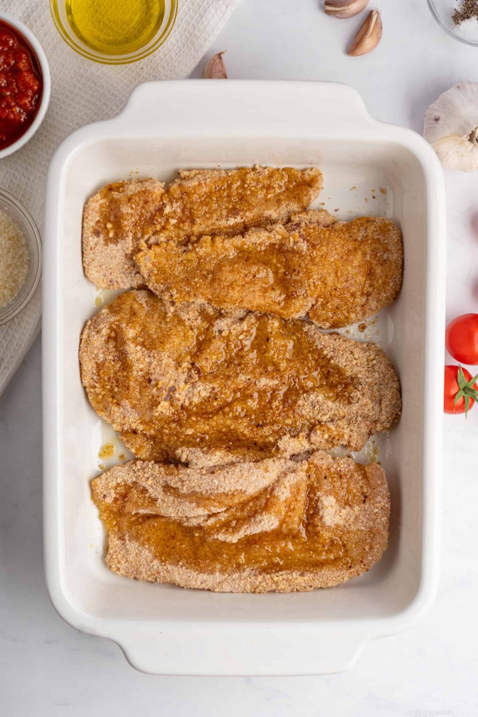 Raw breaded chicken fillets are placed in a white baking dish on a marble surface, ready for cooking. Surrounding the dish are ingredients like olive oil, tomato sauce, garlic, and parmesan cheese on a white towel.