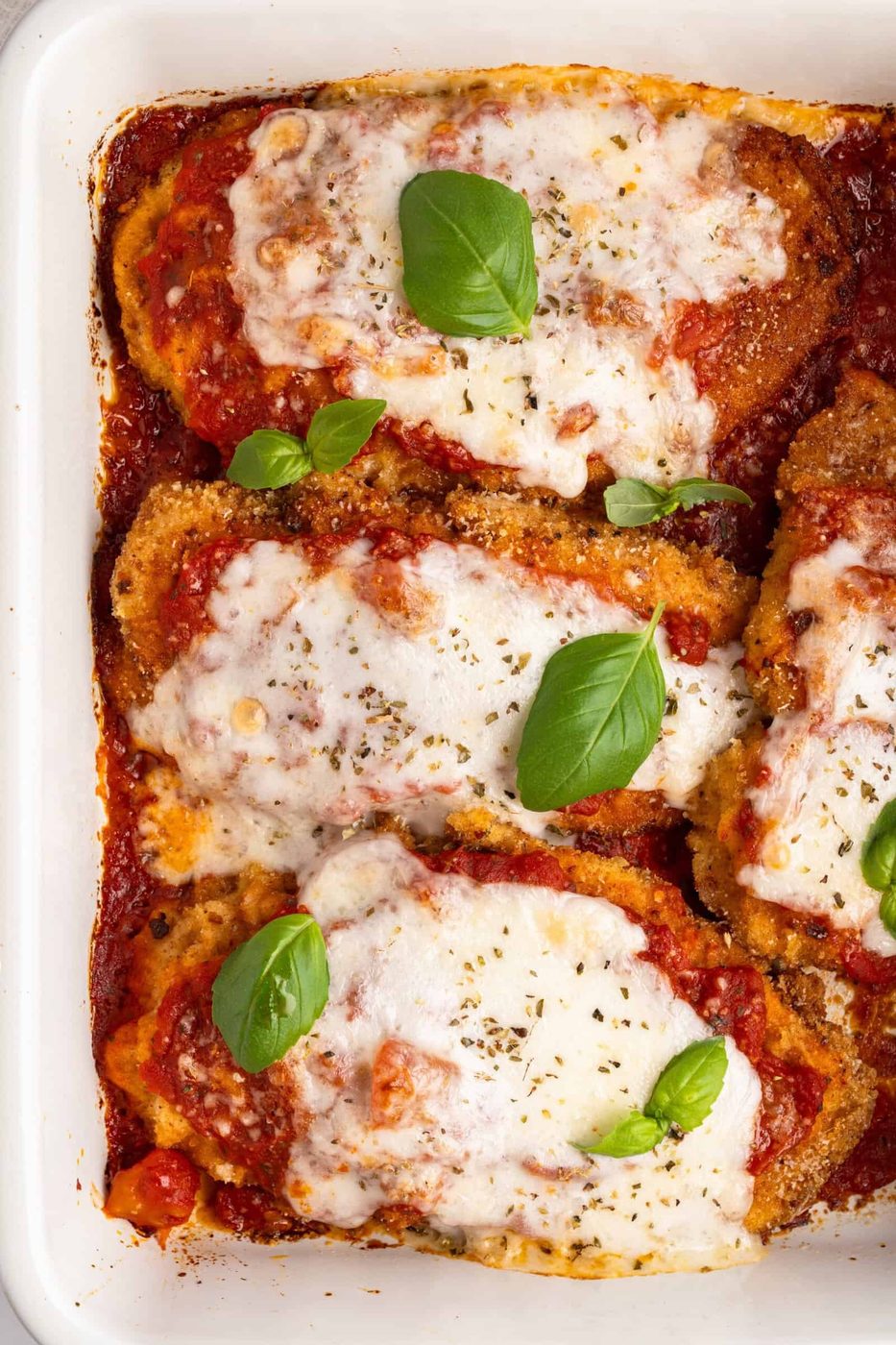 A baking dish with four pieces of chicken parmigiana topped with melted cheese and tomato sauce, garnished with fresh basil leaves. The dish is golden and crispy around the edges.
