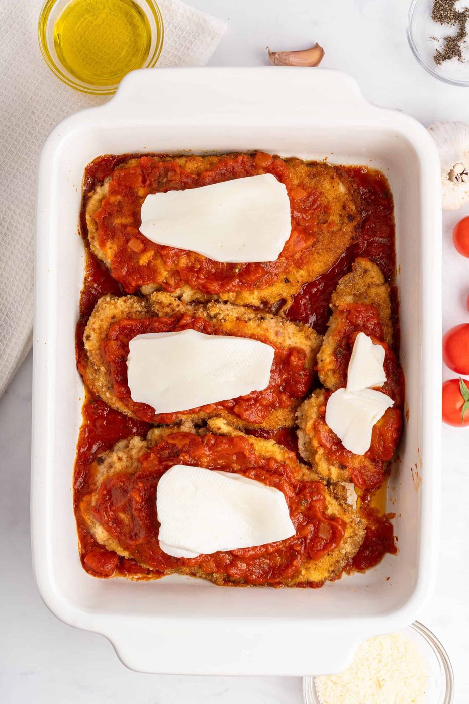 A baking dish containing breaded chicken breasts topped with tomato sauce and slices of cheese. Surrounding the dish are ingredients like tomatoes, garlic, mushrooms, a bowl of grated cheese, and a small dish of olive oil.