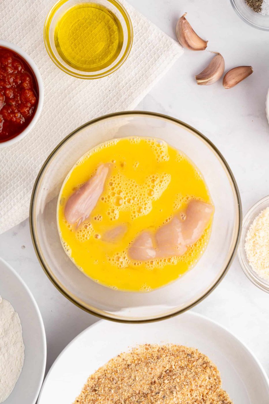 Raw chicken strips are dipped in a bowl of beaten eggs. Surrounding the bowl are ingredients including olive oil, garlic cloves, tomato sauce, flour, breadcrumbs, and seasonings on a white surface.