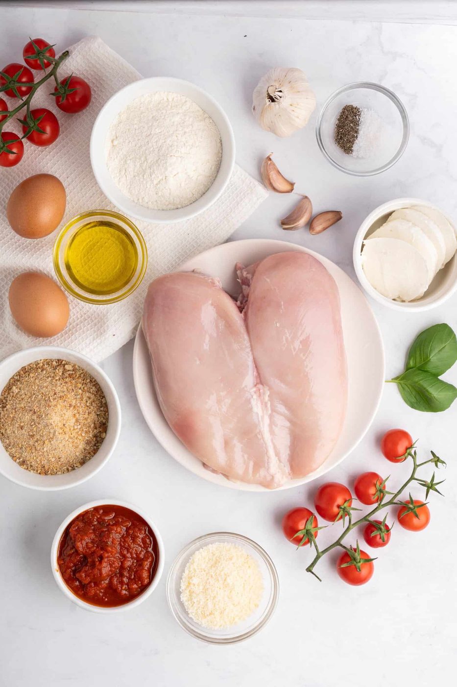 A flatlay of ingredients for a meal: raw chicken breasts, eggs, flour, breadcrumbs, olive oil, marinara sauce, mozzarella, grated cheese, cherry tomatoes, garlic cloves, black pepper, and fresh basil on a white surface.