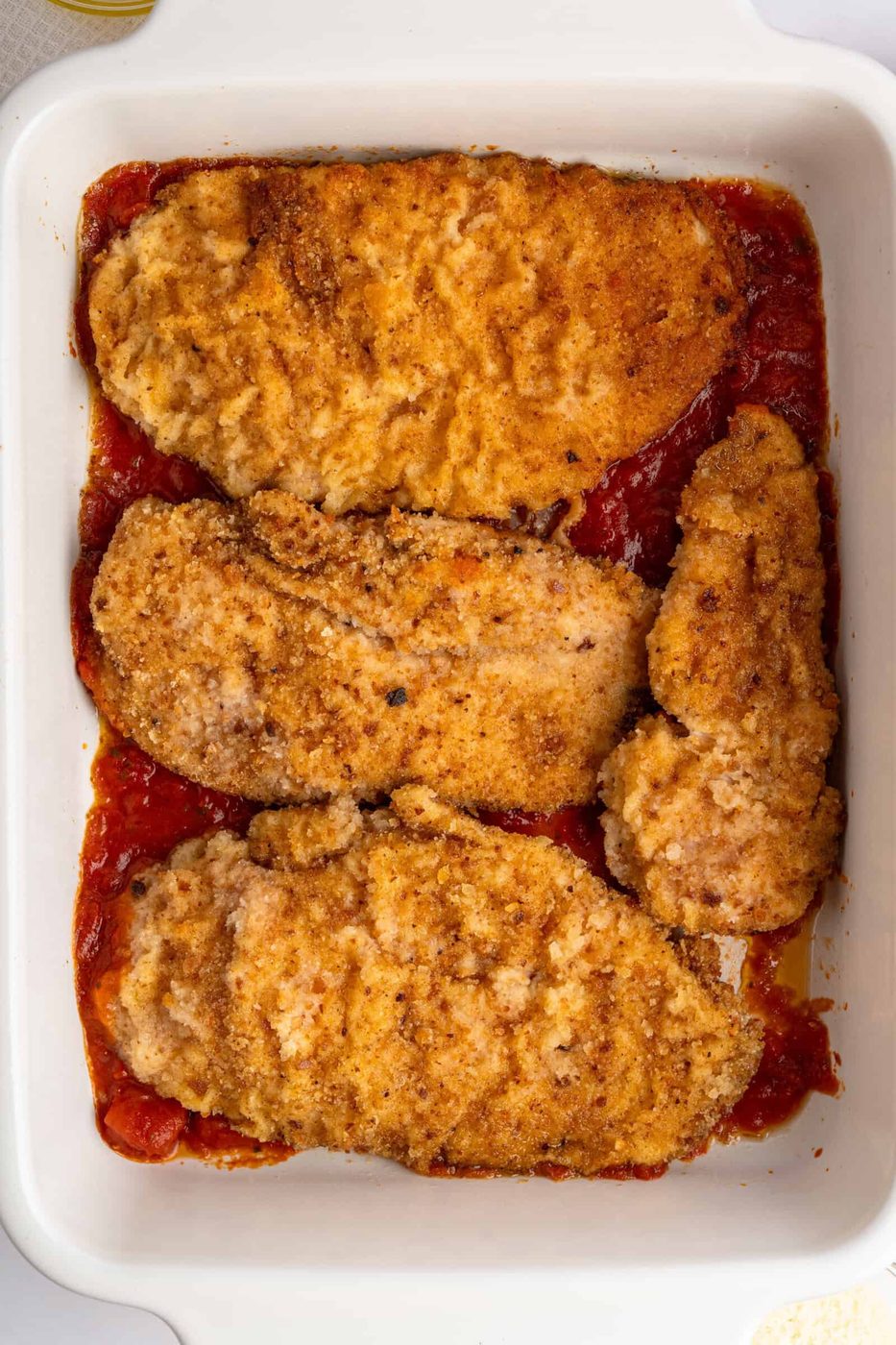 A white baking dish contains four breaded and browned chicken cutlets on a bed of red tomato sauce.