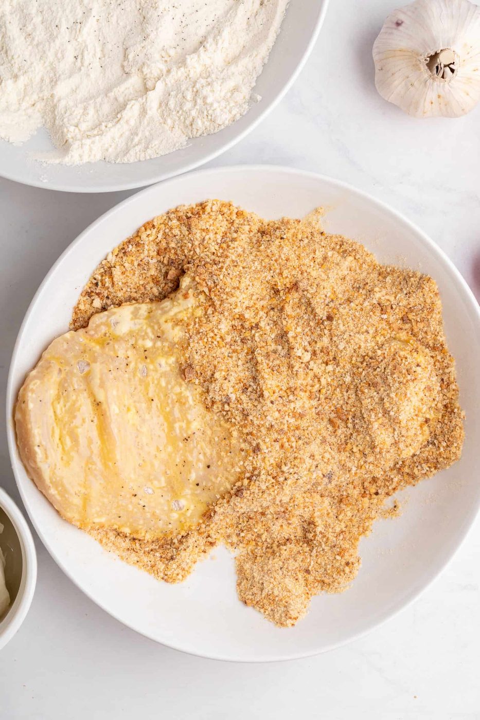 A bowl of chicken breast being coated in seasoned breadcrumbs, with flour and garlic nearby. The chicken appears partially covered, ready for cooking.