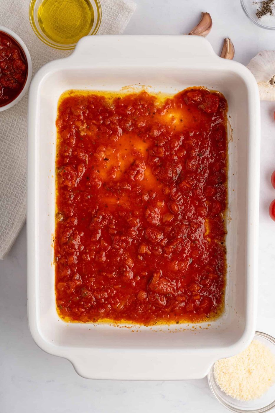 A white rectangular baking dish filled with tomato sauce on a light surface. Nearby are ingredients including olive oil, garlic cloves, and grated cheese, suggesting preparation for a dish.