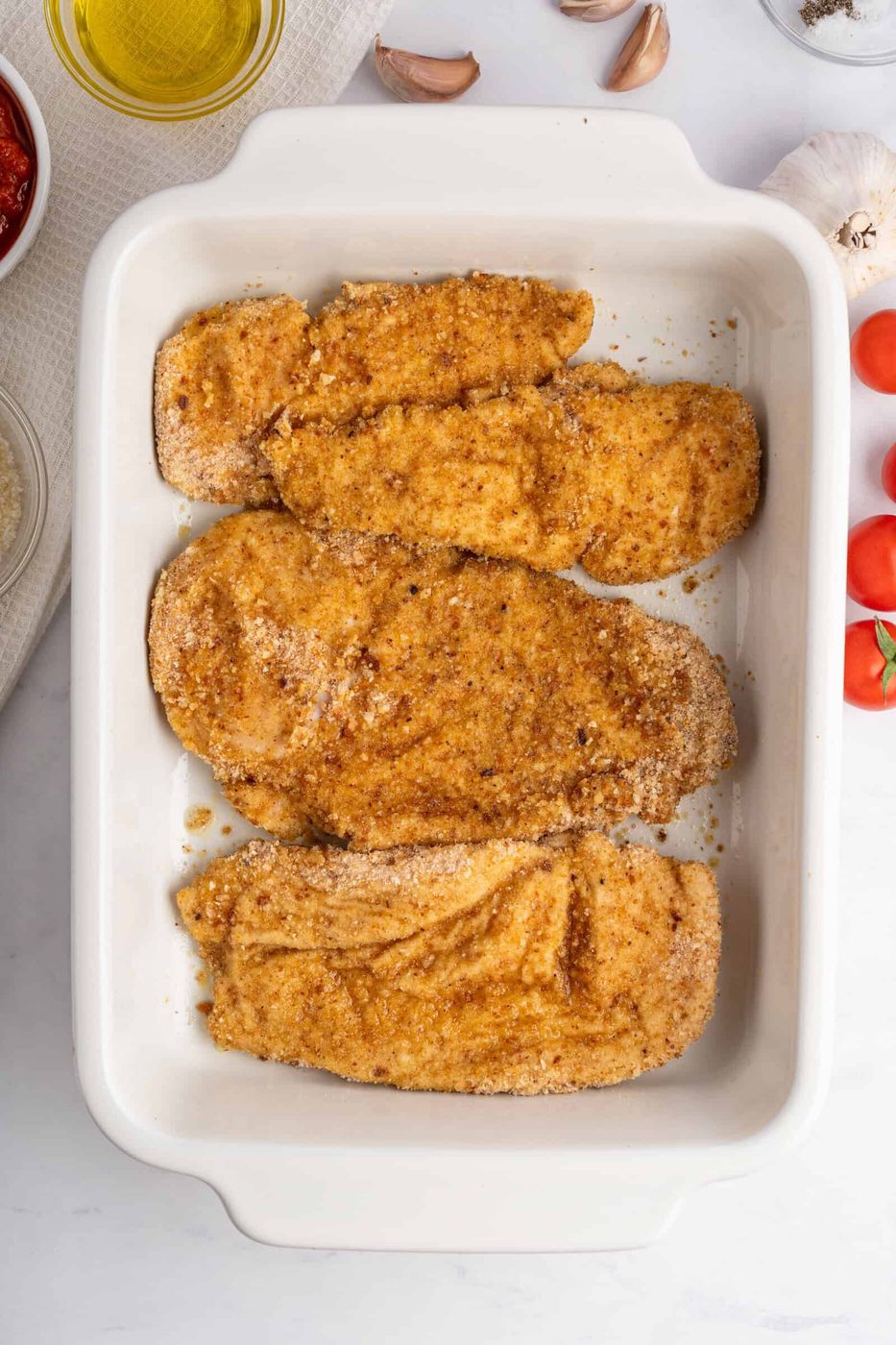 Top view of a white baking dish containing four breaded chicken cutlets. The cutlets are seasoned with spices and breadcrumbs. Ingredients like cherry tomatoes, garlic, and olive oil are nearby on a marble countertop.
