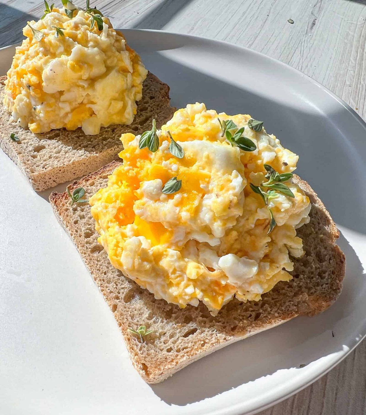 Two slices of whole grain bread topped with scrambled eggs and garnished with fresh herbs, served on a white plate in bright natural light.
