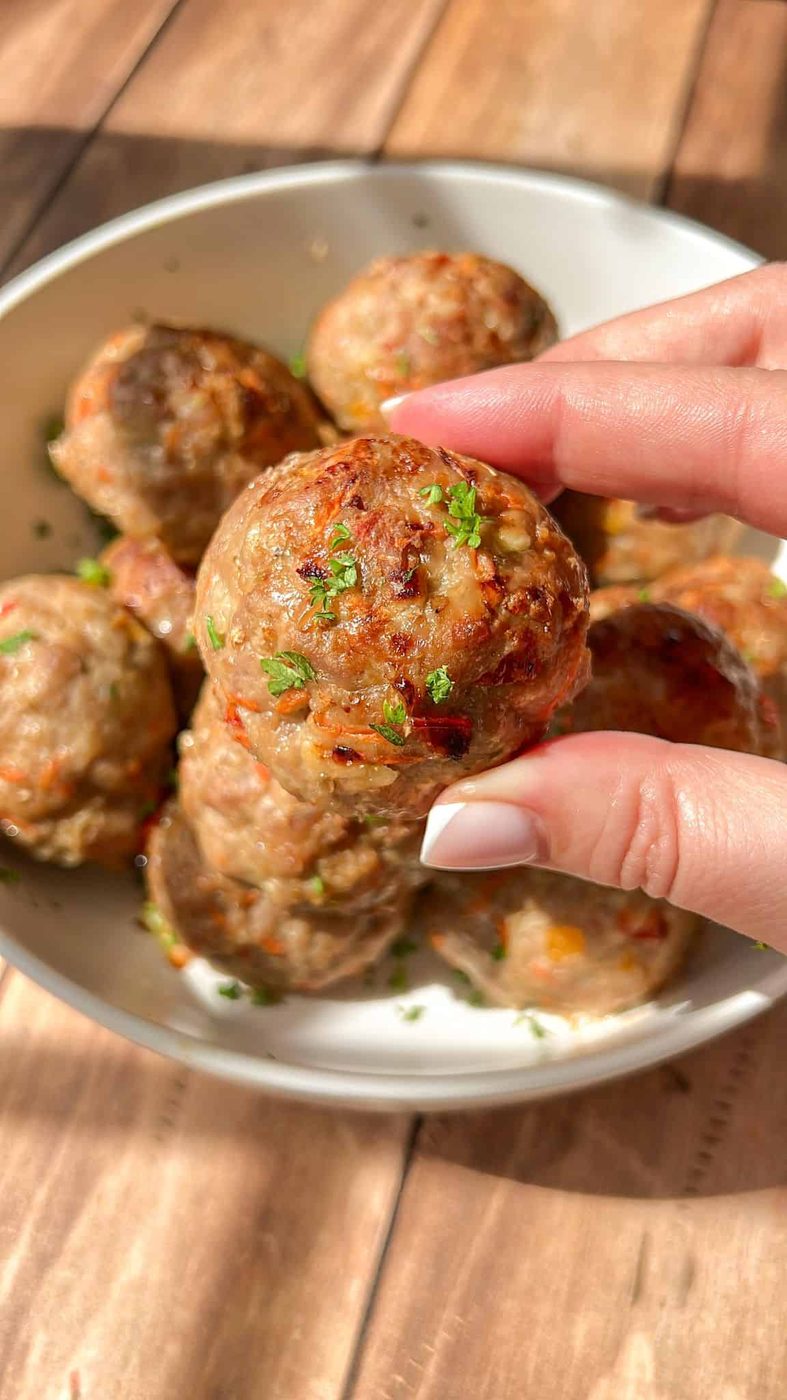 Close-up of a hand holding a cooked meatball garnished with herbs above a bowl filled with more meatballs. The meatballs are on a wooden table, basking in warm natural light.