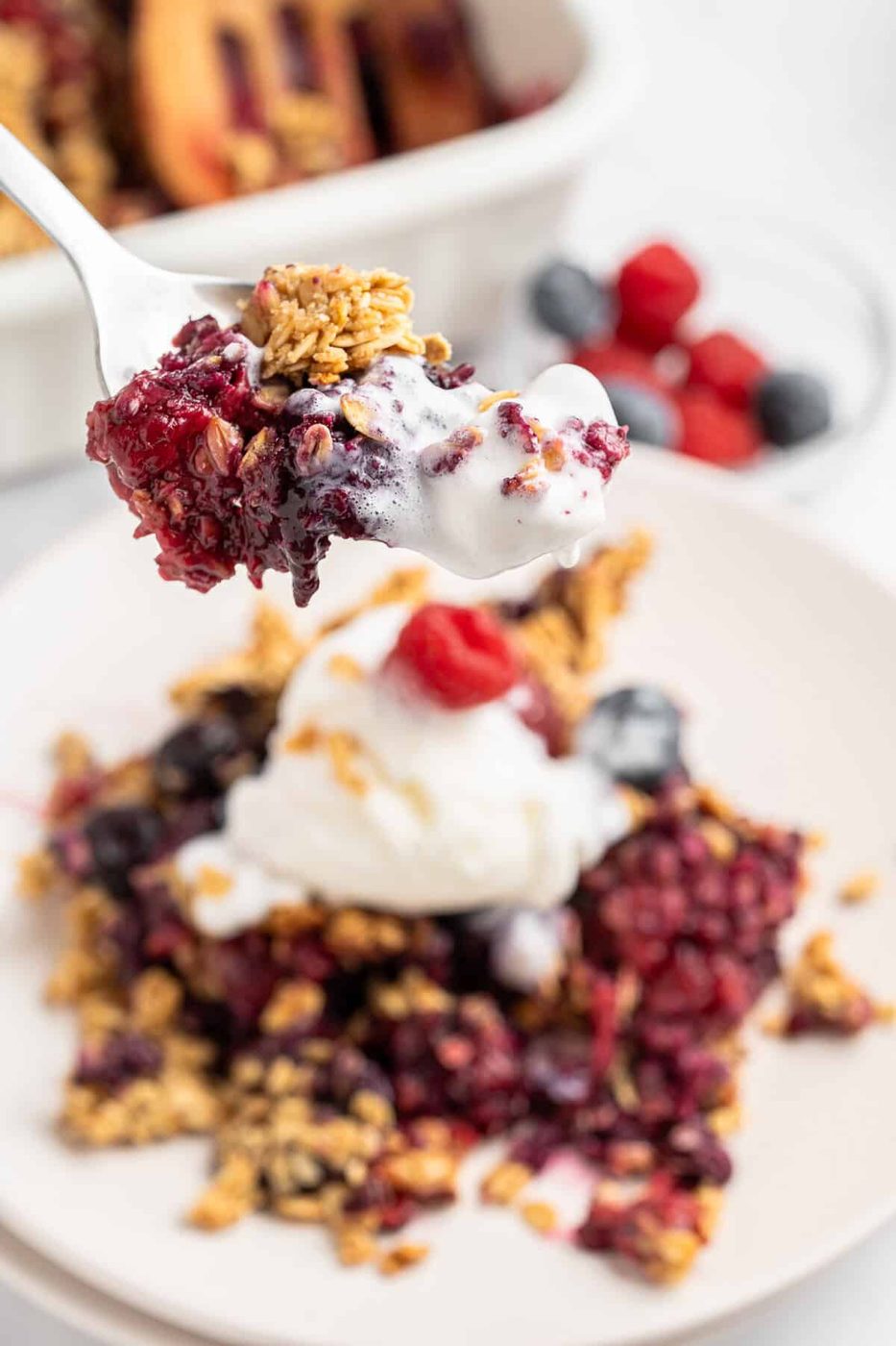 A spoonful of the crisp with granola and a dollop of whipped cream is held above a plate. In the background, a dish with more berry crisp and a small bowl of fresh berries are visible.
