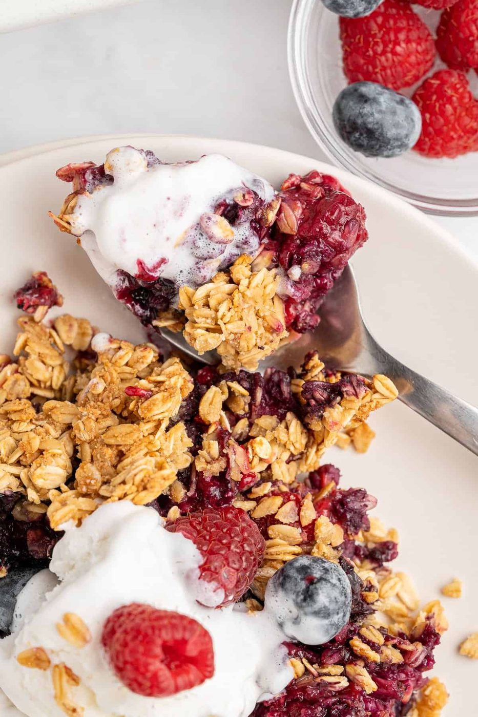 A close-up of a breakfast dish featuring granola, fresh raspberries, and blueberries on a plate. A spoon with some yogurt and granola is beside it. A small bowl with more berries is in the background.