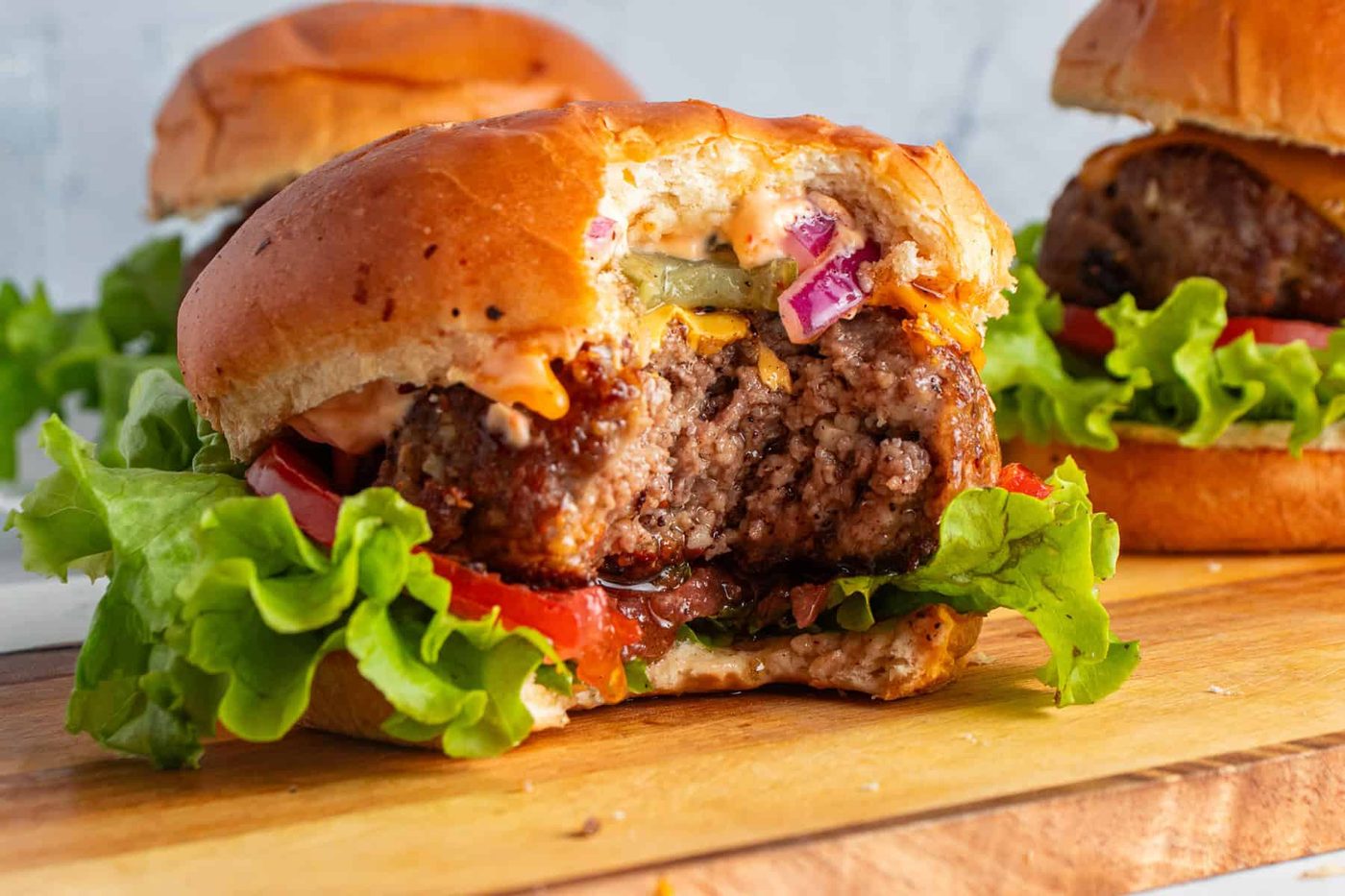 Close-up of a juicy homemade cheeseburger with a bite taken out. It sits on a wooden board, topped with lettuce, tomato, pickles, red onion, and sauce in a soft bun. Other burgers appear blurred in the background.