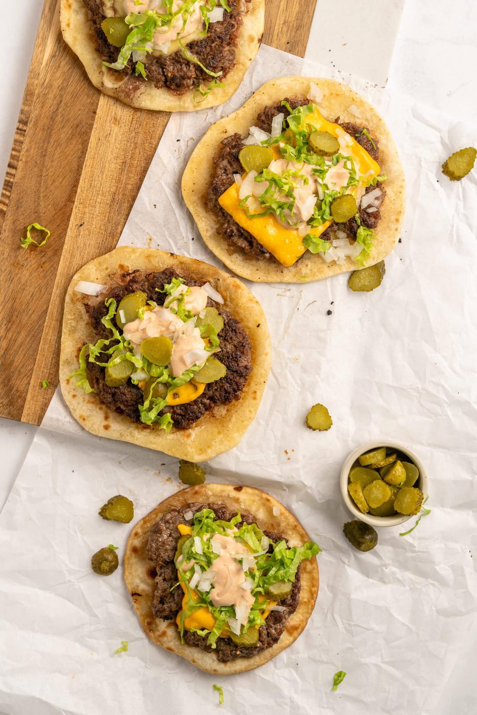 Four flatbread Big Mac smash tacos topped with ground beef, shredded lettuce, pickles, cheese, and a creamy sauce are arranged on parchment paper and a wooden board, with a small bowl of sliced pickles nearby.