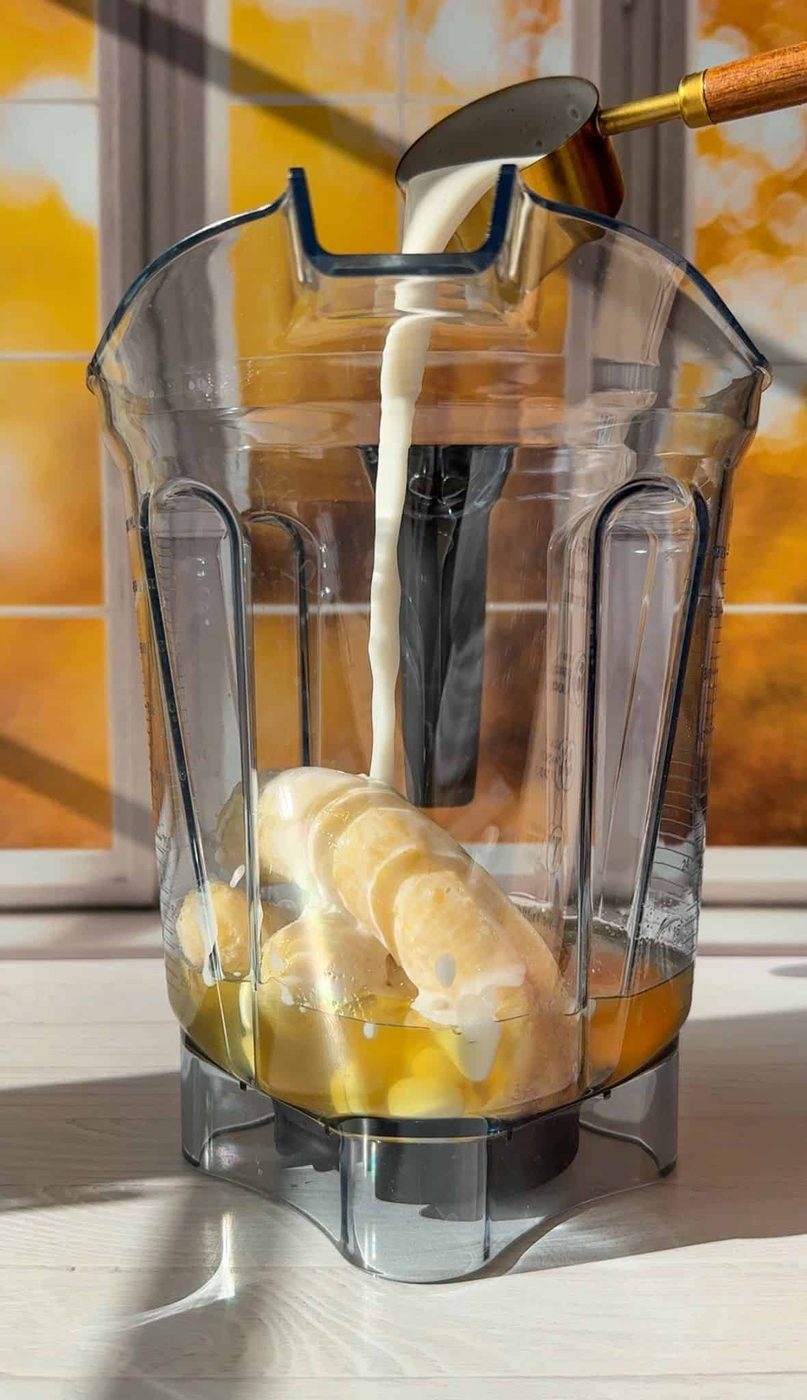 A blender container with bananas, ice, and liquids. Milk is being poured in from a measuring cup. Sunlight shines through a nearby window with yellow foliage visible outside.