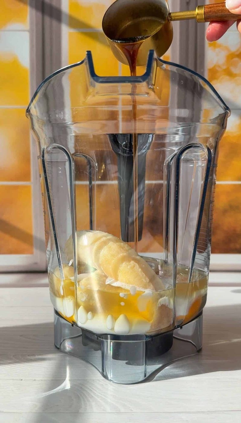 A clear blender pitcher filled with bananas, yogurt, and honey. A hand is pouring a small amount of dark liquid from a spoon into the blender. Sunlight streams in through a window, casting warm light on the scene.