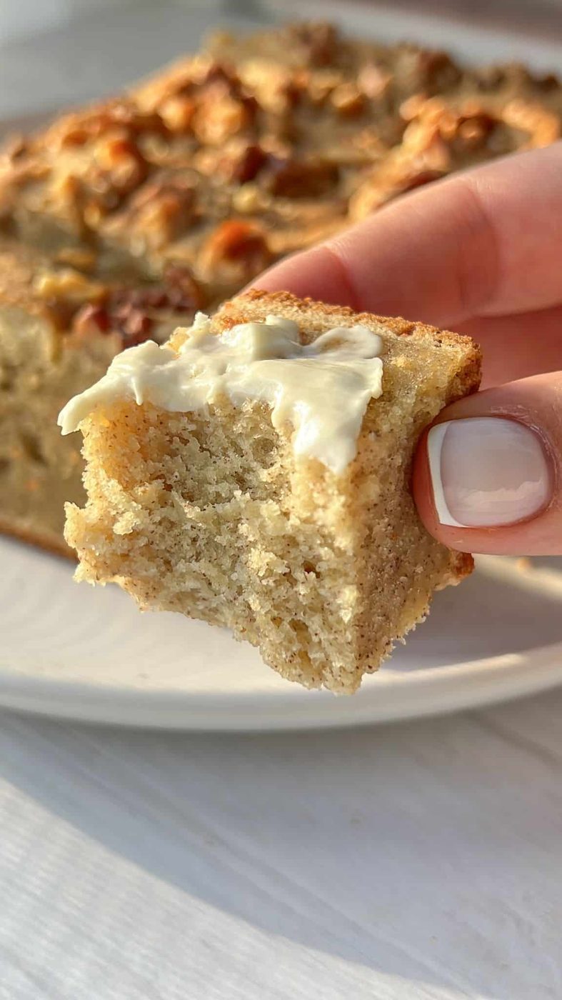 A close-up of a hand holding a piece of banana bread topped with a layer of butter. The bread is moist and studded with nuts, with a bitten section visible, against a blurred background of the whole loaf on a white plate.