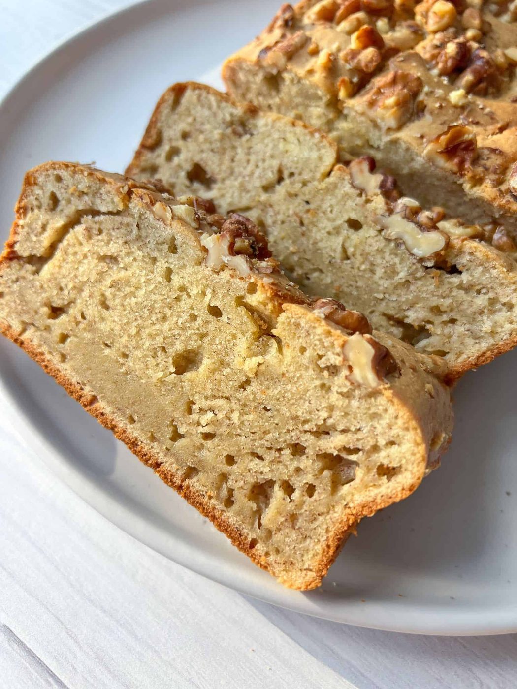 Close-up of sliced banana bread on a white plate. The bread is topped with chopped walnuts, showing its moist texture and golden-brown crust. The scene is set on a light-colored surface, highlighting the bread's appetizing appearance.