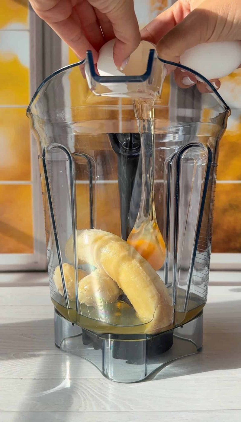 Hands are pouring honey from a bottle into a blender container with bananas inside, preparing ingredients for a smoothie. Sunlight creates a warm glow on the scene.