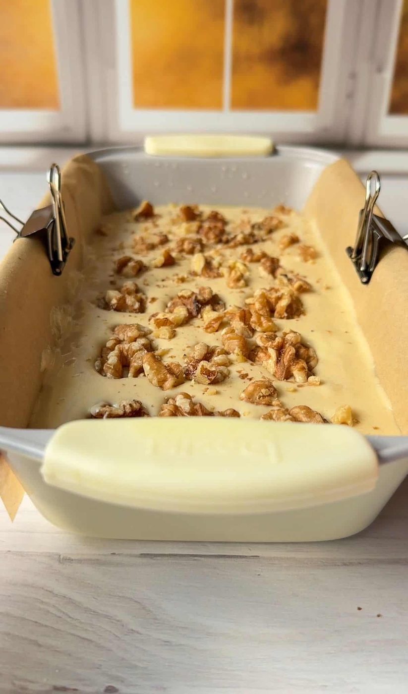 A rectangular baking dish filled with batter topped with chopped walnuts. The dish is lined with parchment paper, held in place by binder clips. A window with a yellow-orange glow is in the background.