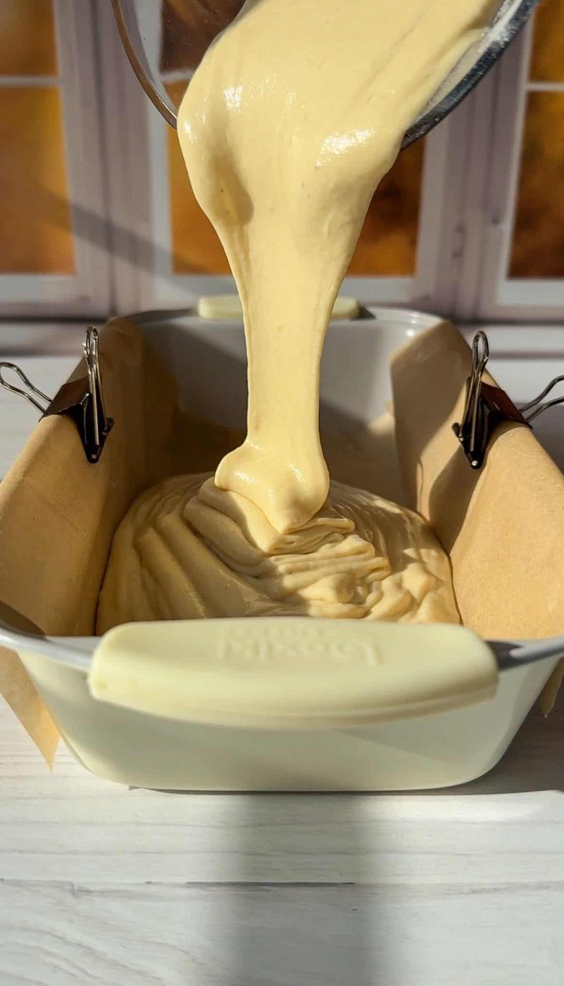Batter is being poured into a parchment-lined loaf pan secured with clips. The scene is bathed in warm sunlight, casting shadows on a white countertop, with a window in the background.