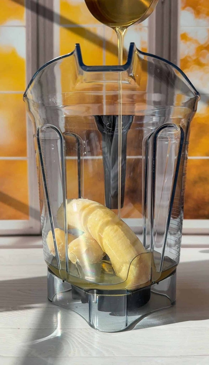 A blender jar with bananas inside sits on a sunlit table. Honey is being poured into the jar from a spoon above. A window with an autumn view is in the background.
