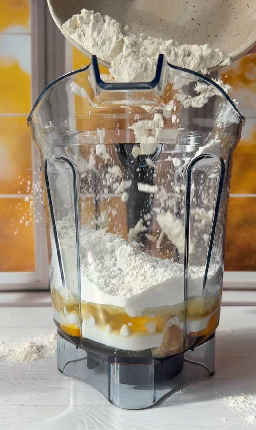 A blender container is being filled with flour from a bowl. The container already holds various baking ingredients, such as eggs, sugar, and milk, on a white countertop, with a sunlit window in the background.