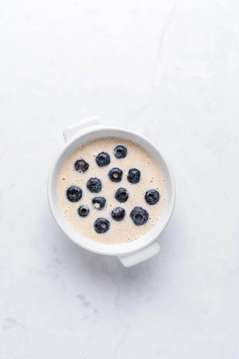 A white bowl filled with a creamy dish topped with scattered blueberries, set on a light-colored marble surface.
