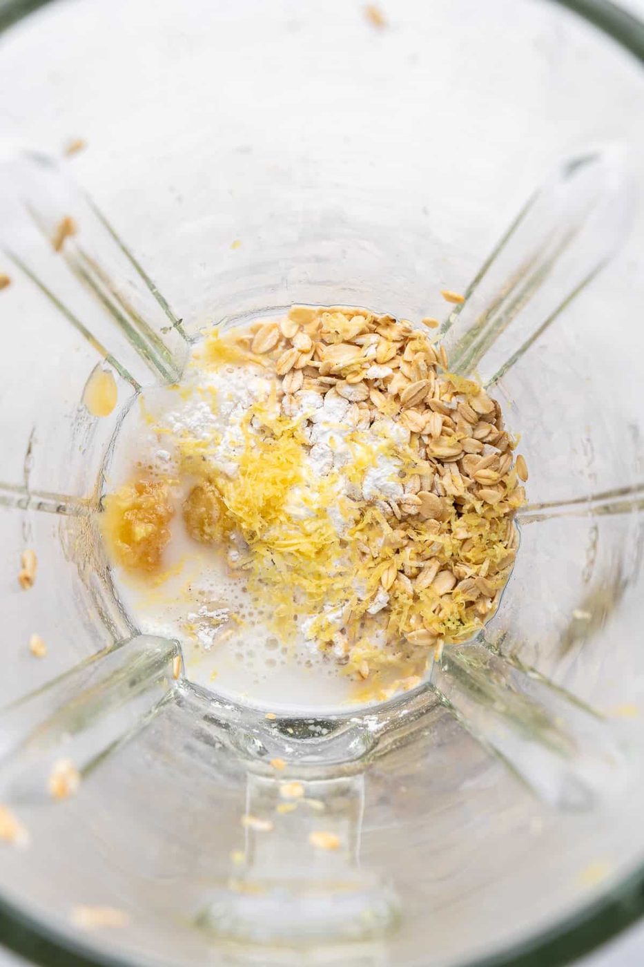 A blender contains oats, lemon zest, milk, and banana. The ingredients are ready to be blended, with oats and lemon zest scattered on top, and milk partially covering them. The mixture appears in a glass blender jug.