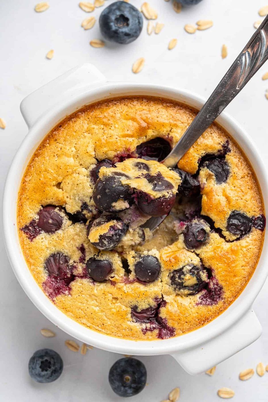 A bowl of baked oatmeal with blueberries on top and scattered around, with a spoon scooping a portion. The dish is set on a light surface with a few oat flakes nearby, creating a cozy breakfast scene.