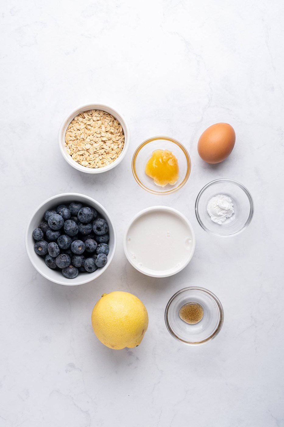 Ingredients for a recipe are arranged on a white marble surface, including a bowl of blueberries, oats, an egg, honey, lemon, milk, baking powder, and vanilla extract.