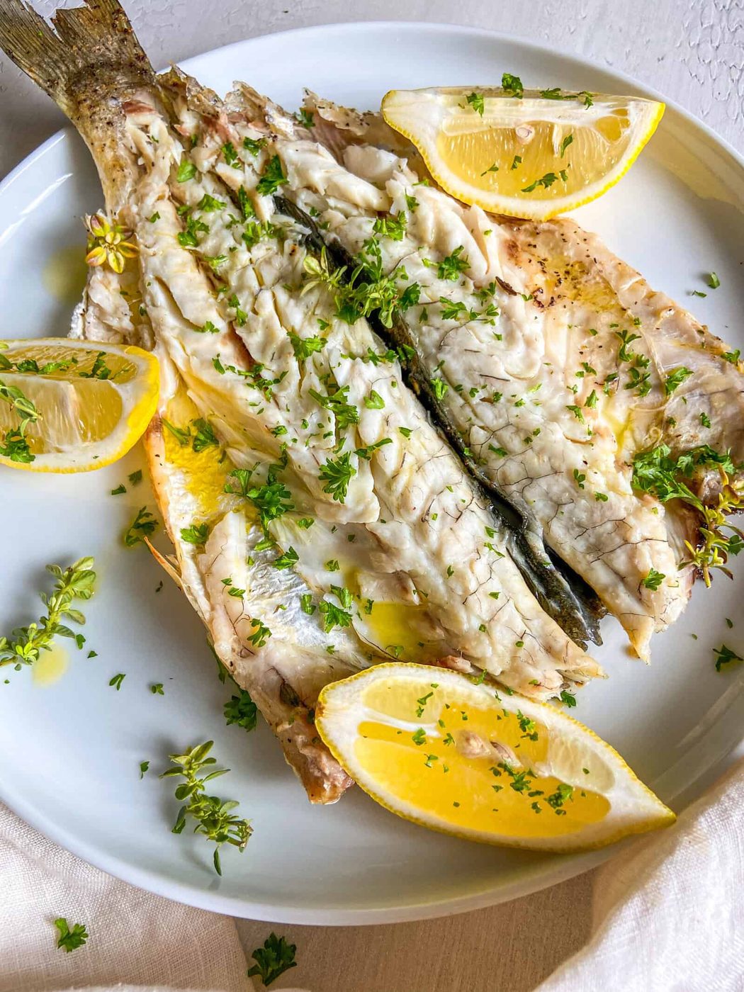 Grilled fish served on a white plate, garnished with chopped parsley and accompanied by lemon wedges. The fish appears tender and flavorful, with a simple yet elegant presentation.