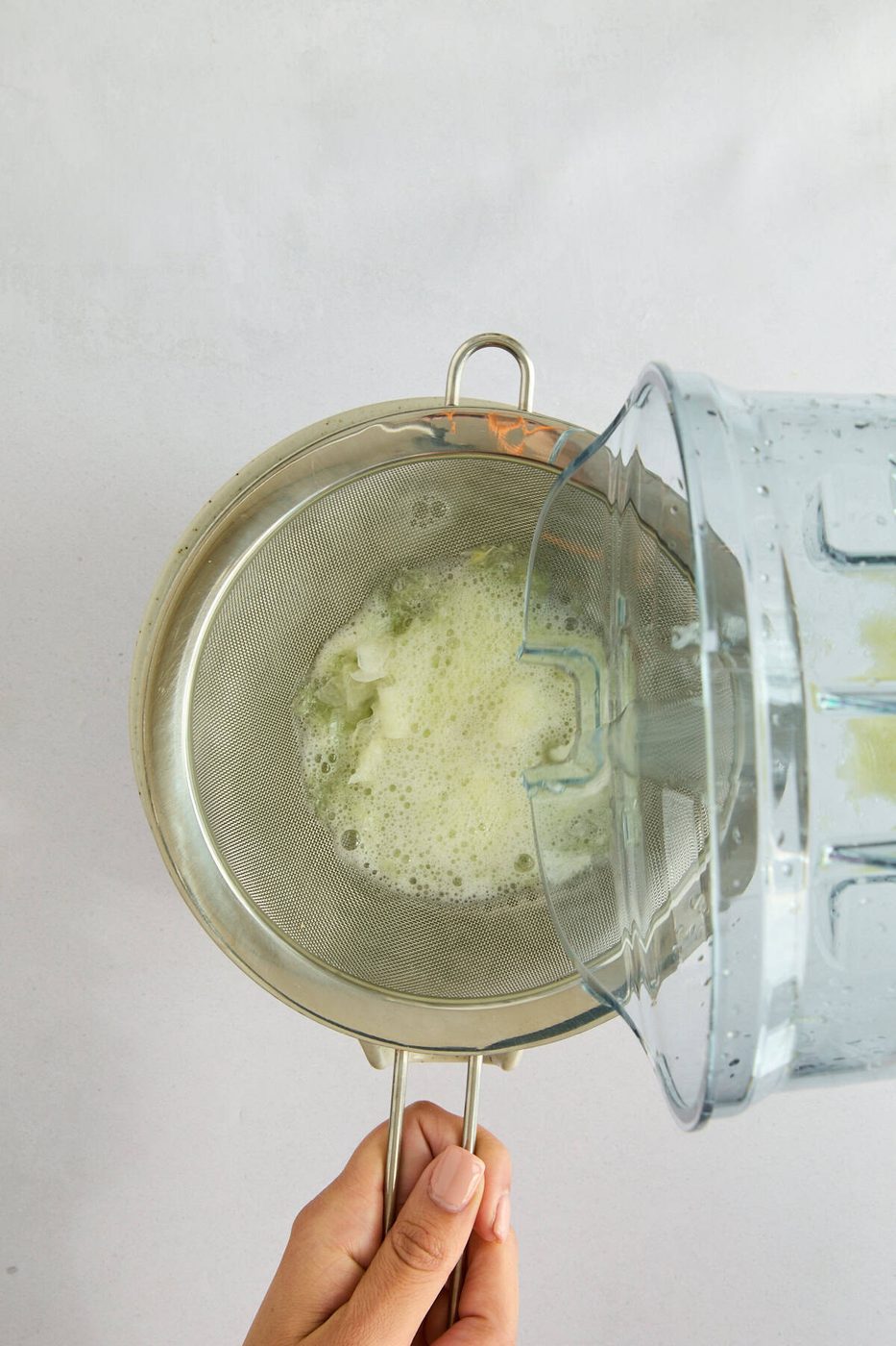 A hand holds a metal strainer over a bowl while pouring a foamy liquid from a blender through the strainer, separating pulp from juice.