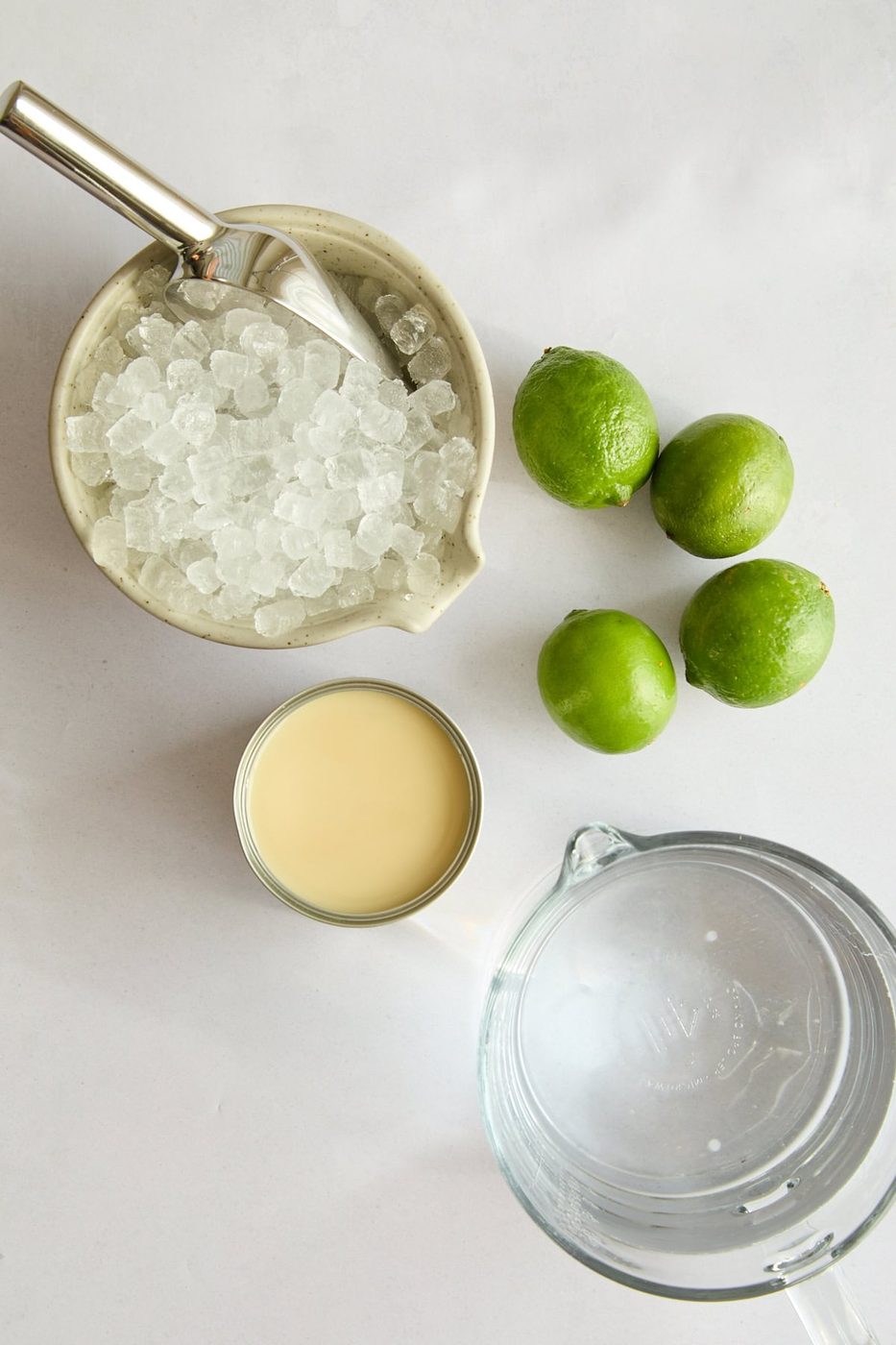 A top-down view of a bowl of crushed ice with a scoop, three whole limes, an open can of sweetened condensed milk, and a glass pitcher of water on a white surface.