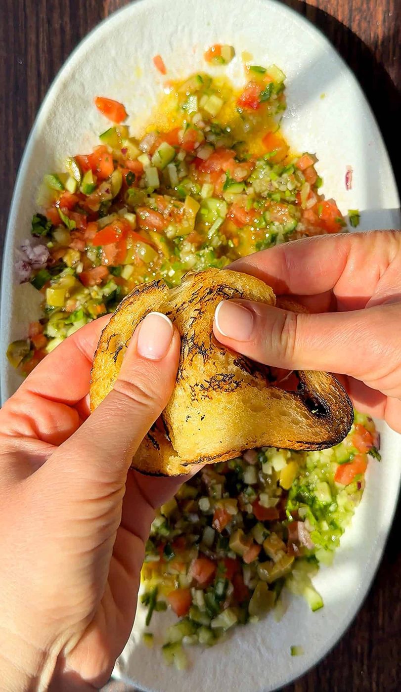 Two hands hold and pinch a charred tortilla chip above a plate of colorful, finely chopped salsa—perfect for pairing with an olive oil bread dip or your favorite bread dipping oil recipe on a white dish.