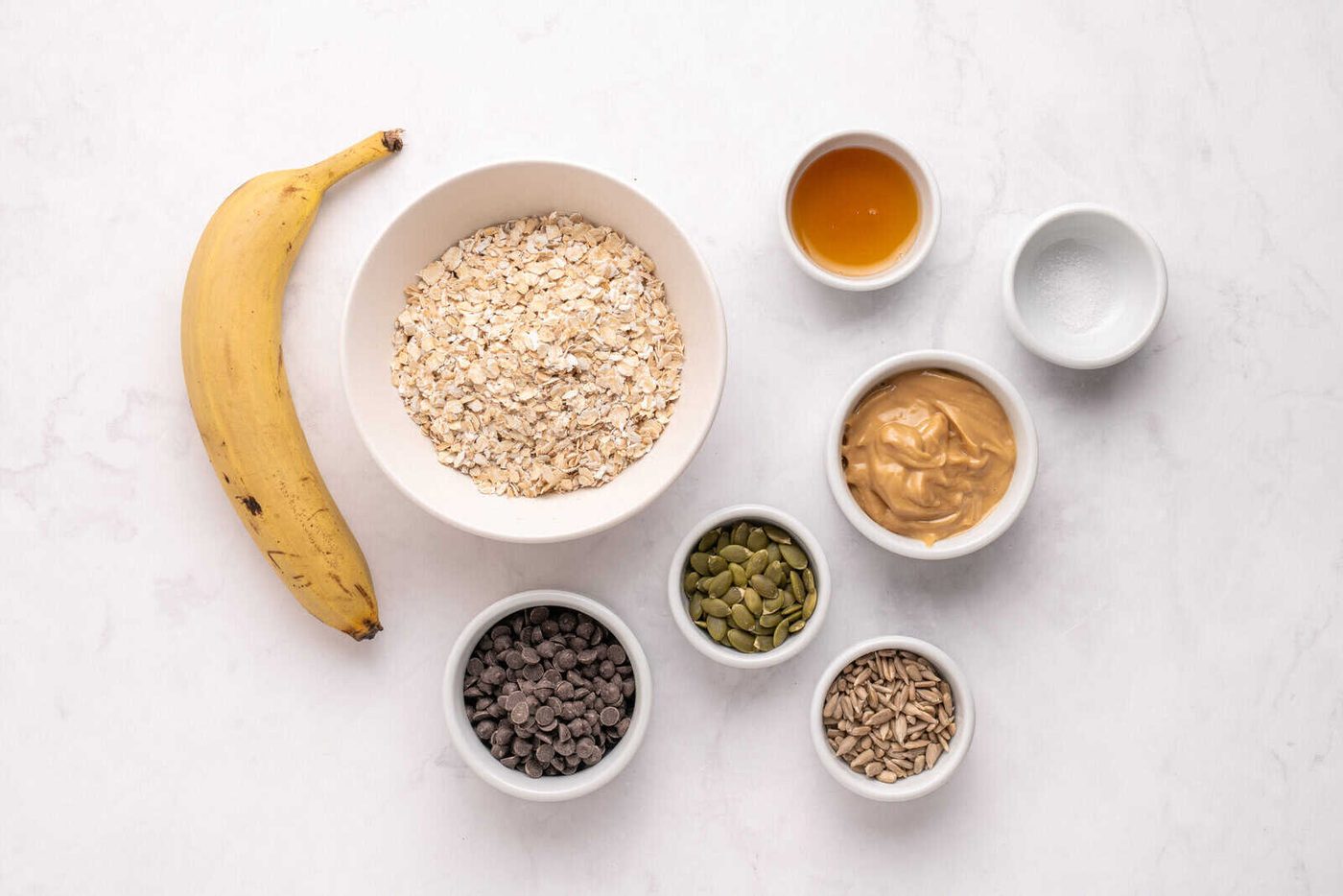 A banana, a bowl of rolled oats, and small bowls of honey, peanut butter, salt, pumpkin seeds, chocolate chips, and sunflower seeds are arranged on a white marble surface.