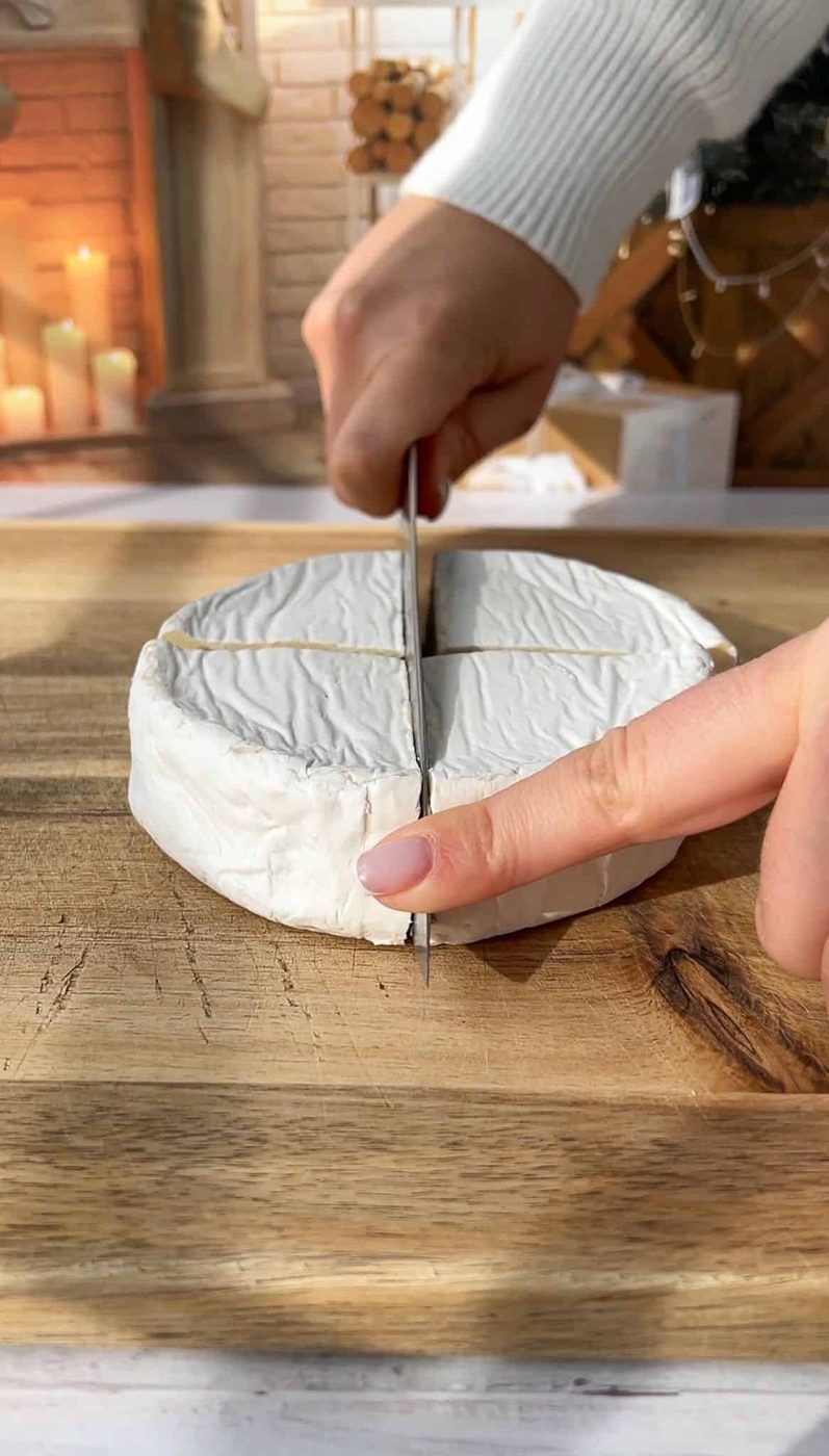 A person slicing a round wheel of brie cheese on a wooden cutting board. The background includes lit candles and stacked firewood, creating a warm and cozy atmosphere.