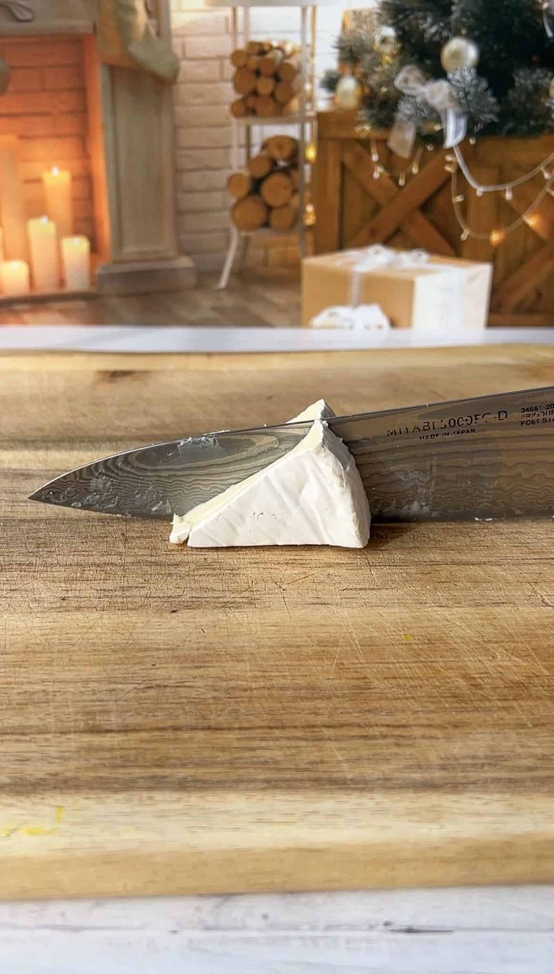 A knife slices through a piece of brie cheese on a wooden cutting board. The background features a cozy, festive room with candles, a fireplace, and holiday decorations.