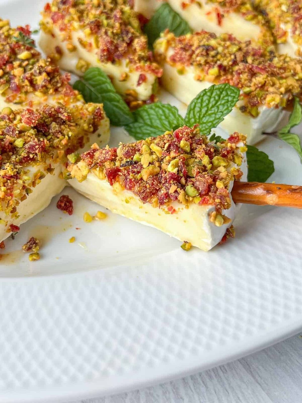 Slices of creamy cheese topped with crushed pistachios and dried fruit are arranged on a white plate. Fresh mint leaves are scattered for garnish, and a pretzel stick is used to pick up one of the slices.