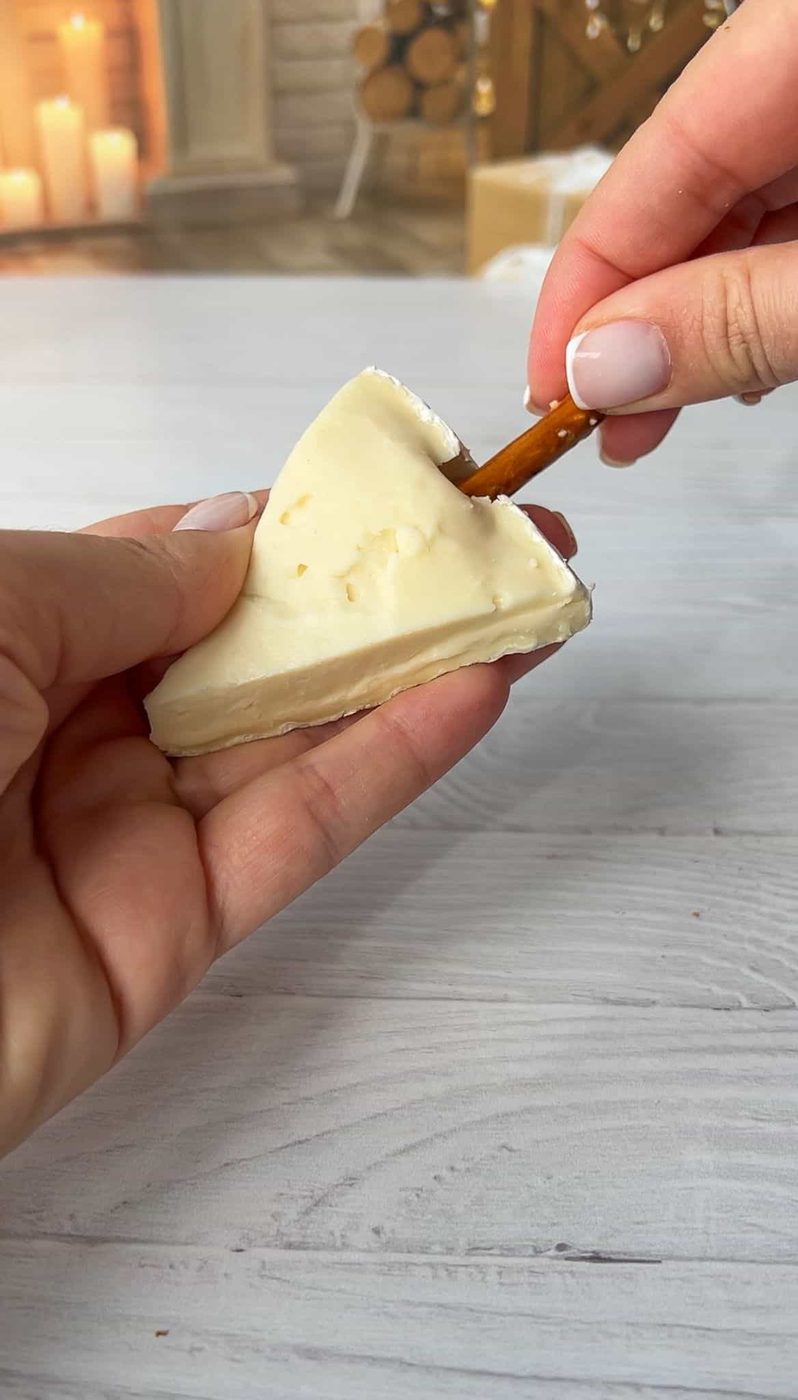 A person holds a wedge of soft cheese with one hand and inserts a pretzel stick into it with the other hand. There are candles and stacked wood in the blurred background, suggesting a cozy setting.
