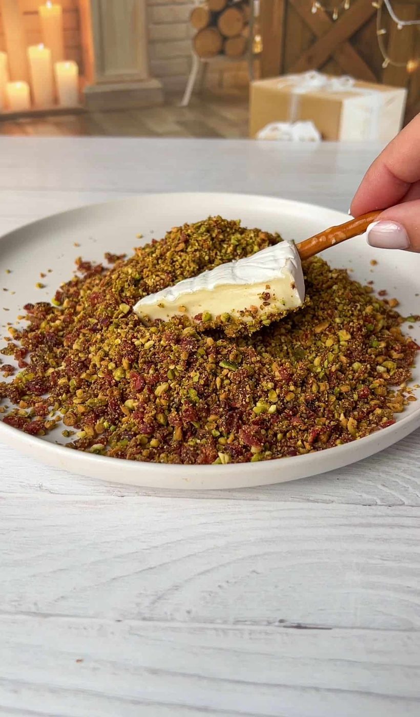 A wedge is being coated with a mixture of finely chopped nuts and herbs on a plate. In the background, there is a cozy setting with candles, logs, and a wrapped gift. 