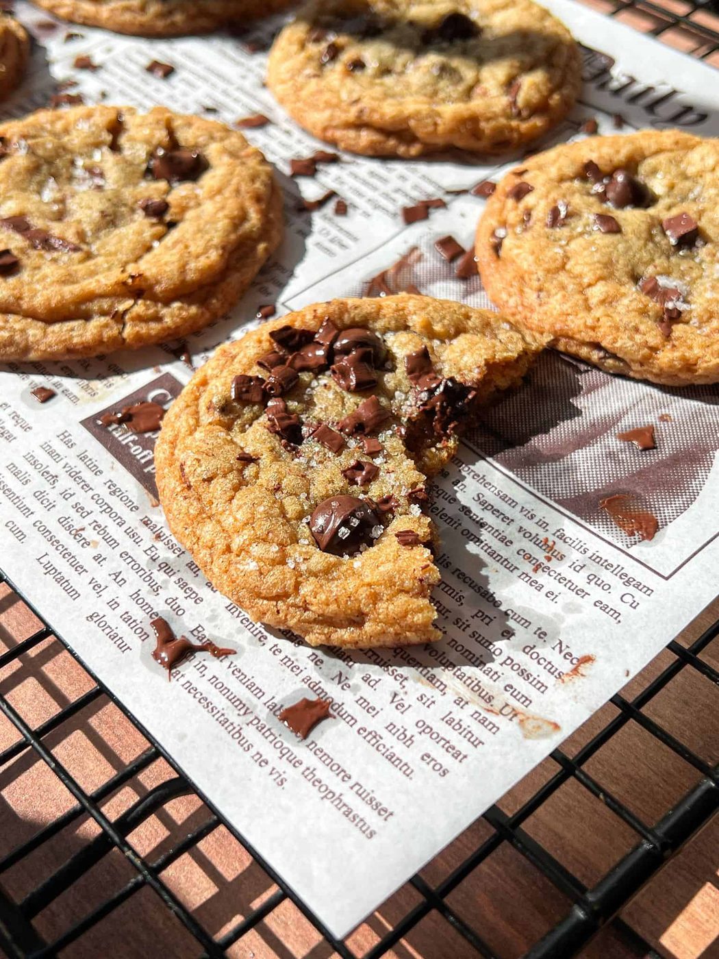 A close-up of Brown Butter Chocolate Chip Cookies on a cooling rack lined with newspaper; one cookie has a large bite taken out, revealing gooey melted chocolate chips inside.