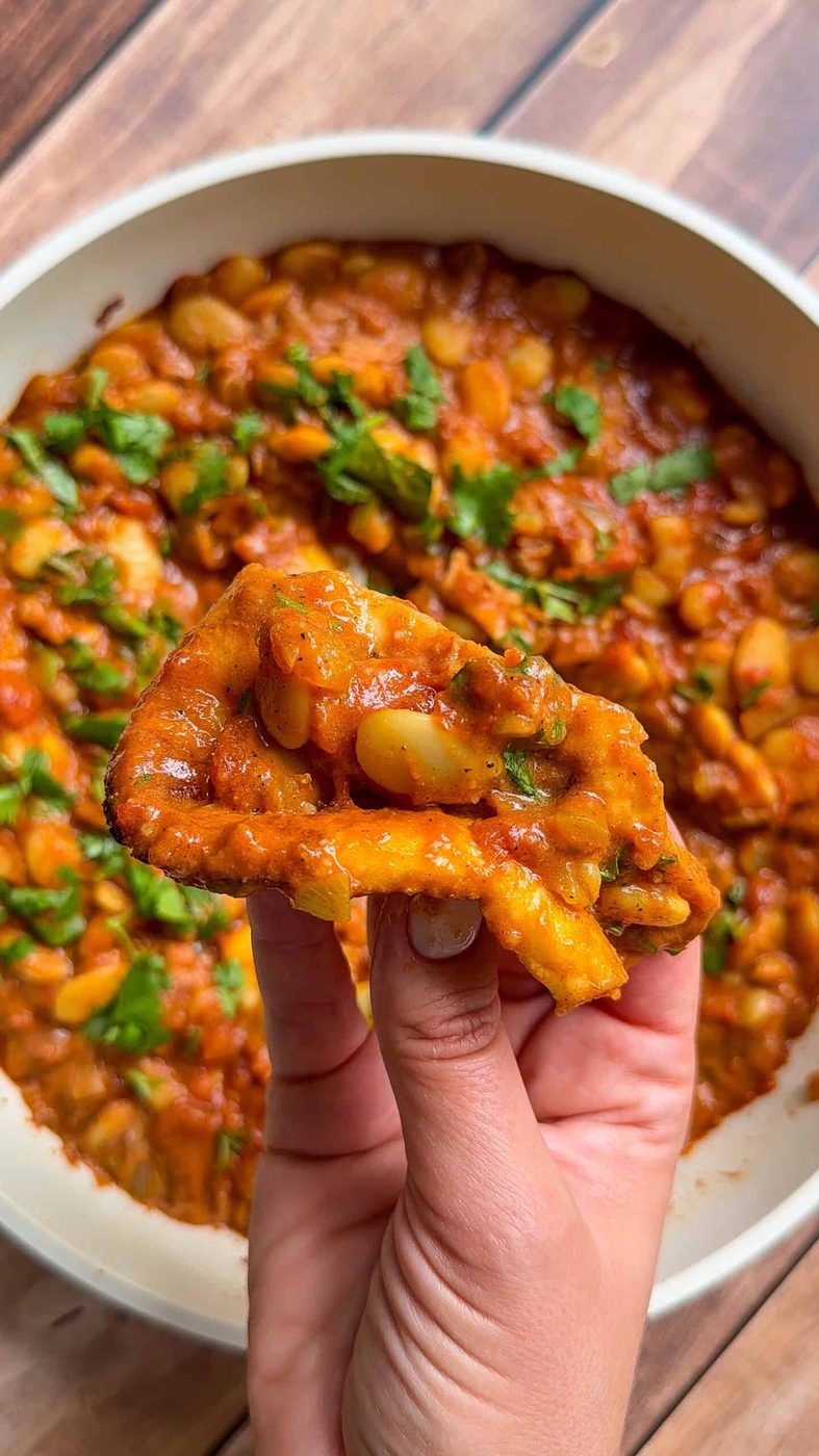 Hand holding fried bread dipped in butter chicken beans