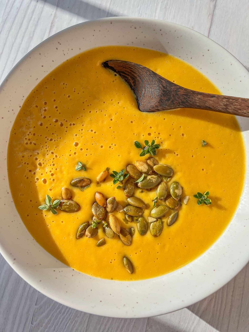 A bowl of creamy Butternut Squash Soup garnished with pumpkin seeds and fresh thyme, with a wooden spoon resting inside. The soup is placed on a light-colored wooden surface, with sunlight casting soft shadows.
