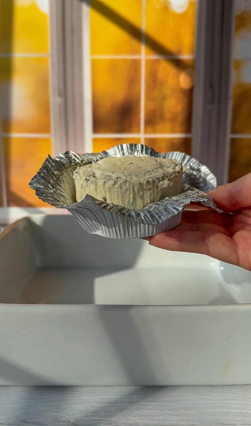 A hand holding an opened foil-wrapped round of cheese over a white dish. Warm sunlight streams through a window with a grid pattern in the background.