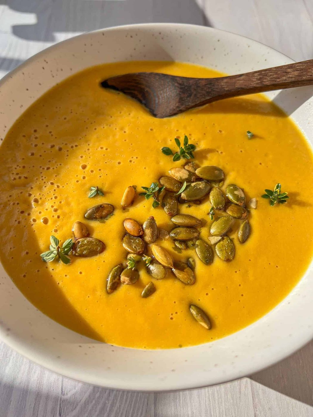 A creamy bowl of Butternut Squash Soup garnished with pumpkin seeds and fresh thyme, served in a white bowl with a wooden spoon. Sunlight casts a natural shadow on the light-colored surface.