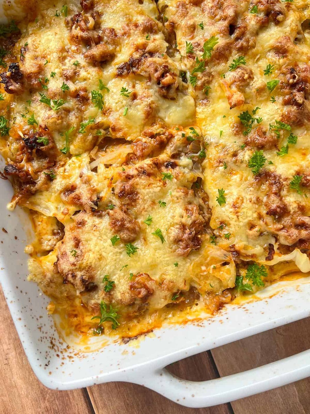 A close-up of a Cabbage Lasagna casserole in a white dish, topped with melted cheese, browned ground meat, and sprinkled with fresh chopped parsley on a wooden surface.